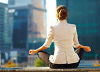 Woman in business clothes meditating, city buildings in the background.