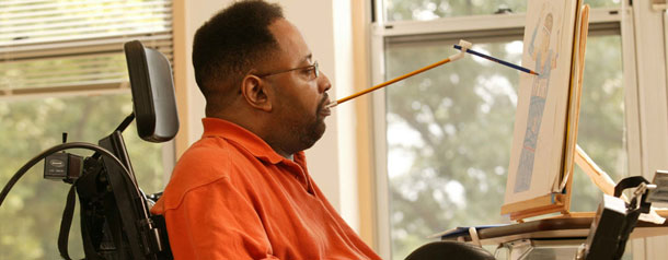 A man in a wheelchair unable to use his hands drawing a picture using a mouth stick  
