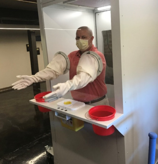 Photo of a man in an enclosed booth with his hands and arms encased in protective gloves. There is a shelf capable of holding supplies connected to the outside of the booth.